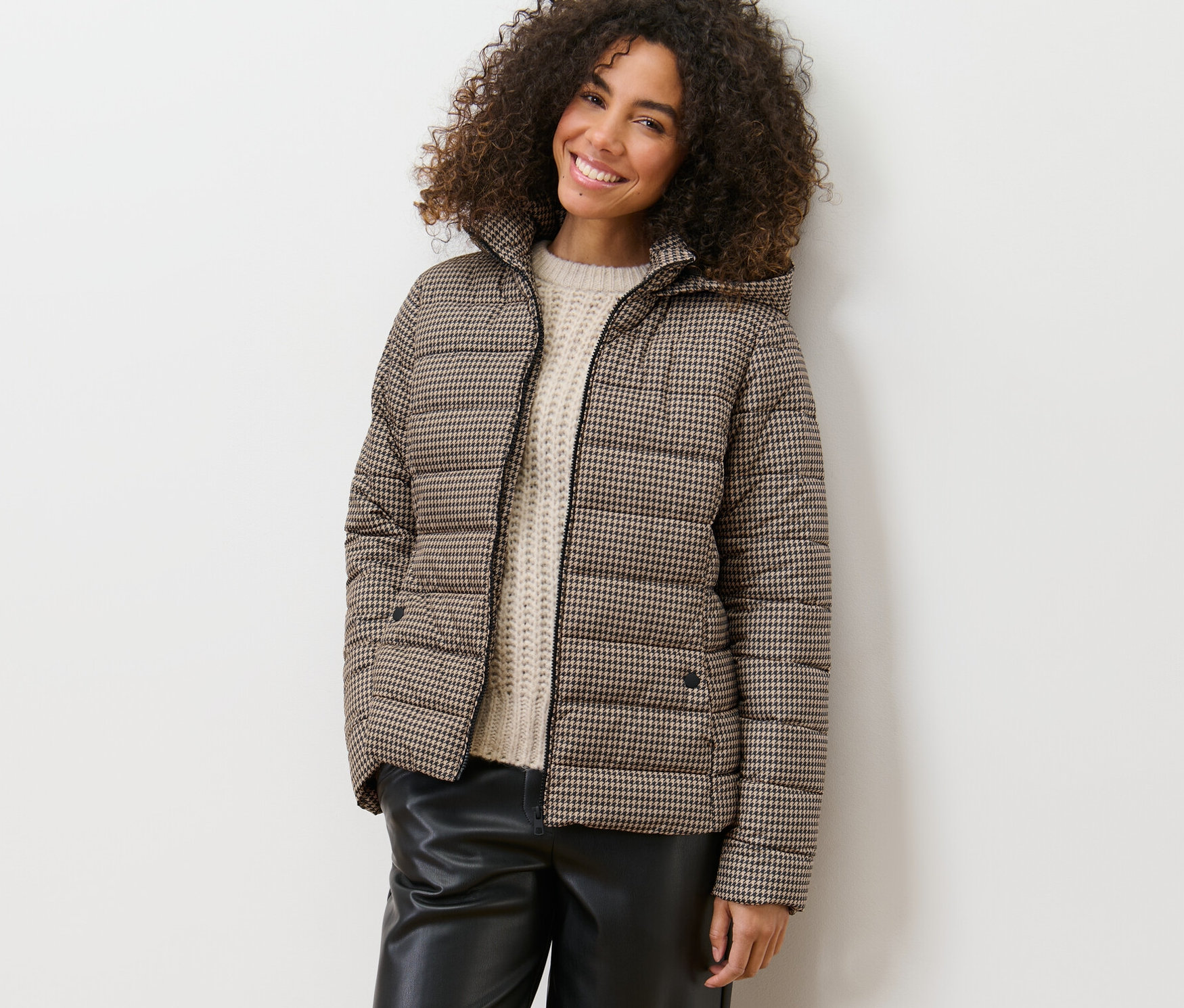 Eine Frau lächelt und lehnt an einer Wand in einer bedruckten Steppjacke mit Kapuze.