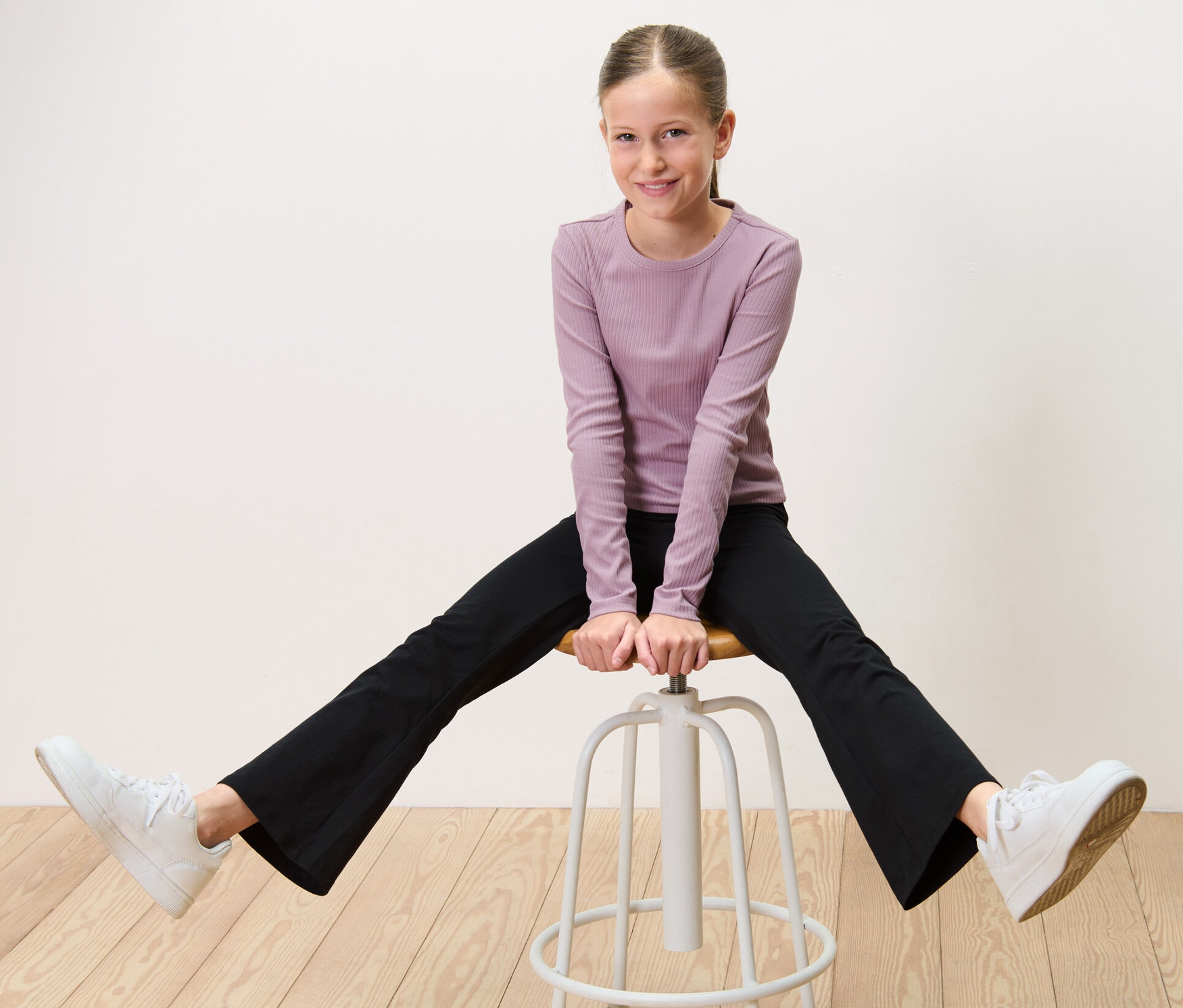 Mädchen sitzt breitbeinig auf einem Hocker und trägt 2 Kinder-Flared-Leggings.