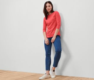 Frau posiert in einem korallenfarbenen Shirt mit 3/4-Arm, blauen Jeans und weißen Turnschuhen.