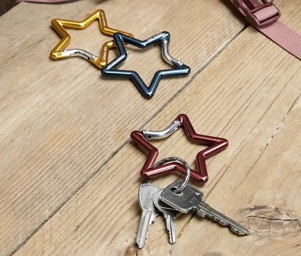 Drei sternförmige Karabinerhaken in Gold, Blau und Rot liegen auf einem Holztisch, wobei an dem roten Karabinerhaken Schlüssel befestigt sind. Im Hintergrund ist ein Teil eines Rucksacks zu sehen.