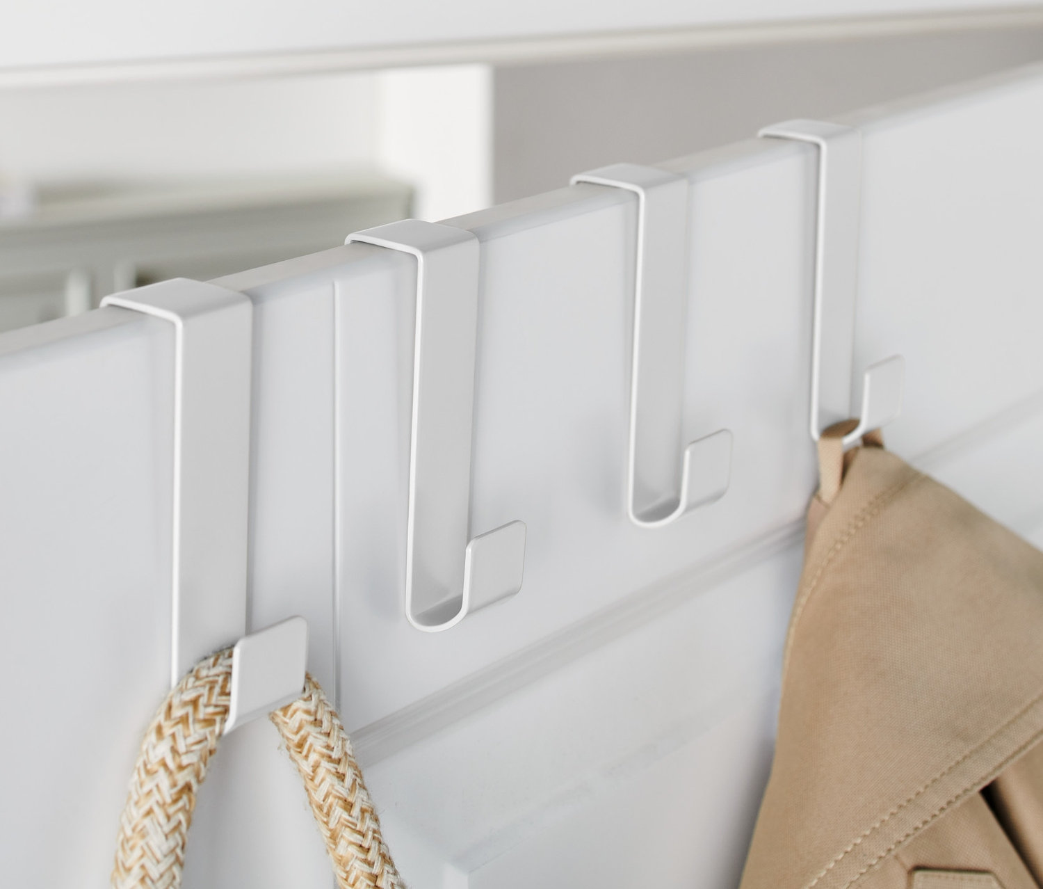Drei weiße Türhaken hängen an einer weißen Tür, an denen eine Jacke und eine Tasche hängen.
