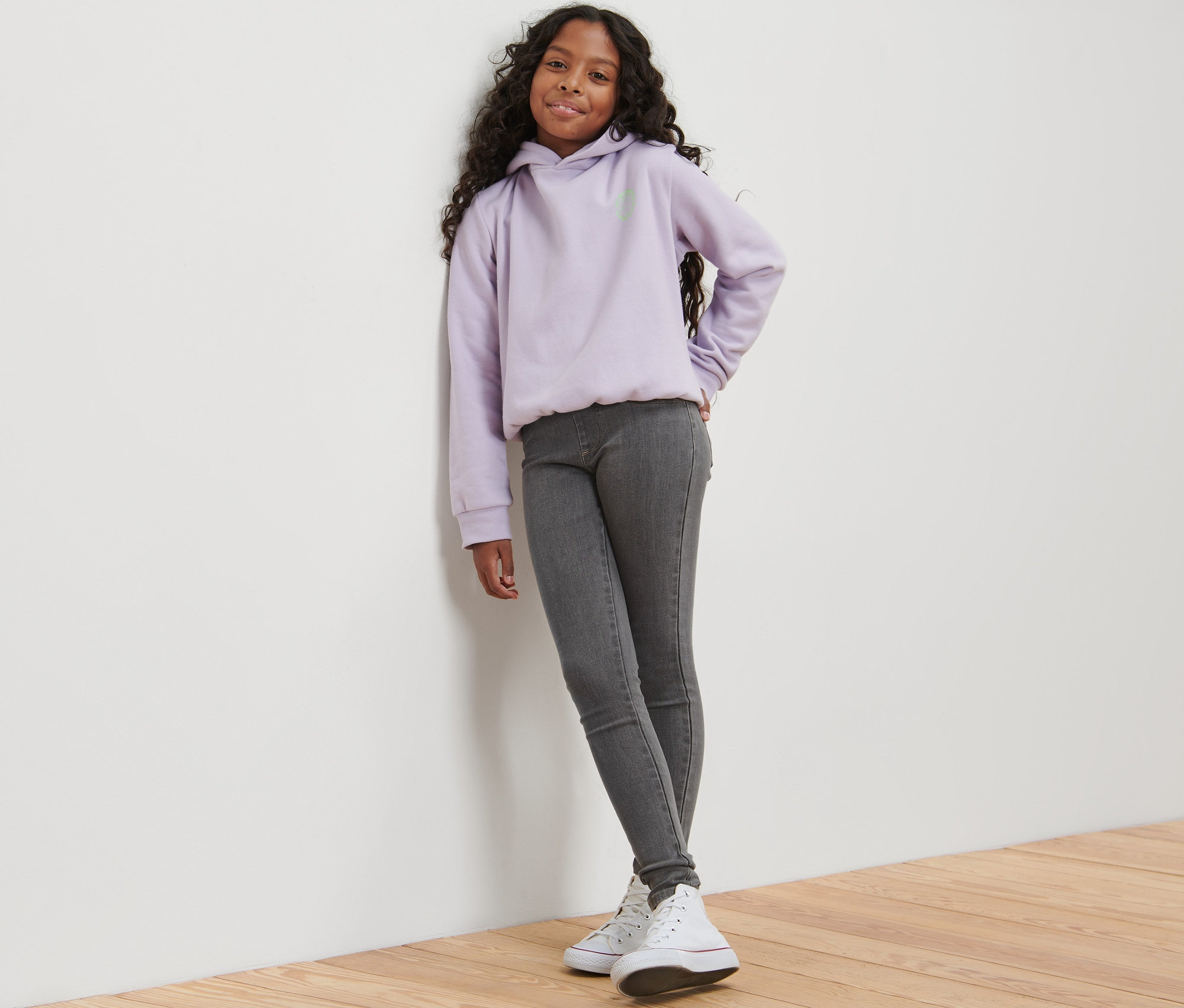 Mädchen lehnt an Wand, trägt graue Kinder-Treggings – Fit »Jule« und lila Hoodie.