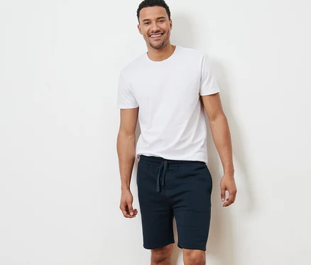 Ein lächelnder Mann steht vor einer weißen Wand, trägt ein weißes T-Shirt und Sweatshorts.