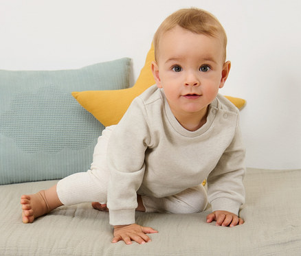 Ein Baby krabbelt auf einer Unterlage neben Kissen. Es trägt 2 Baby-Sweatshirts mit angerauter Innenseite.