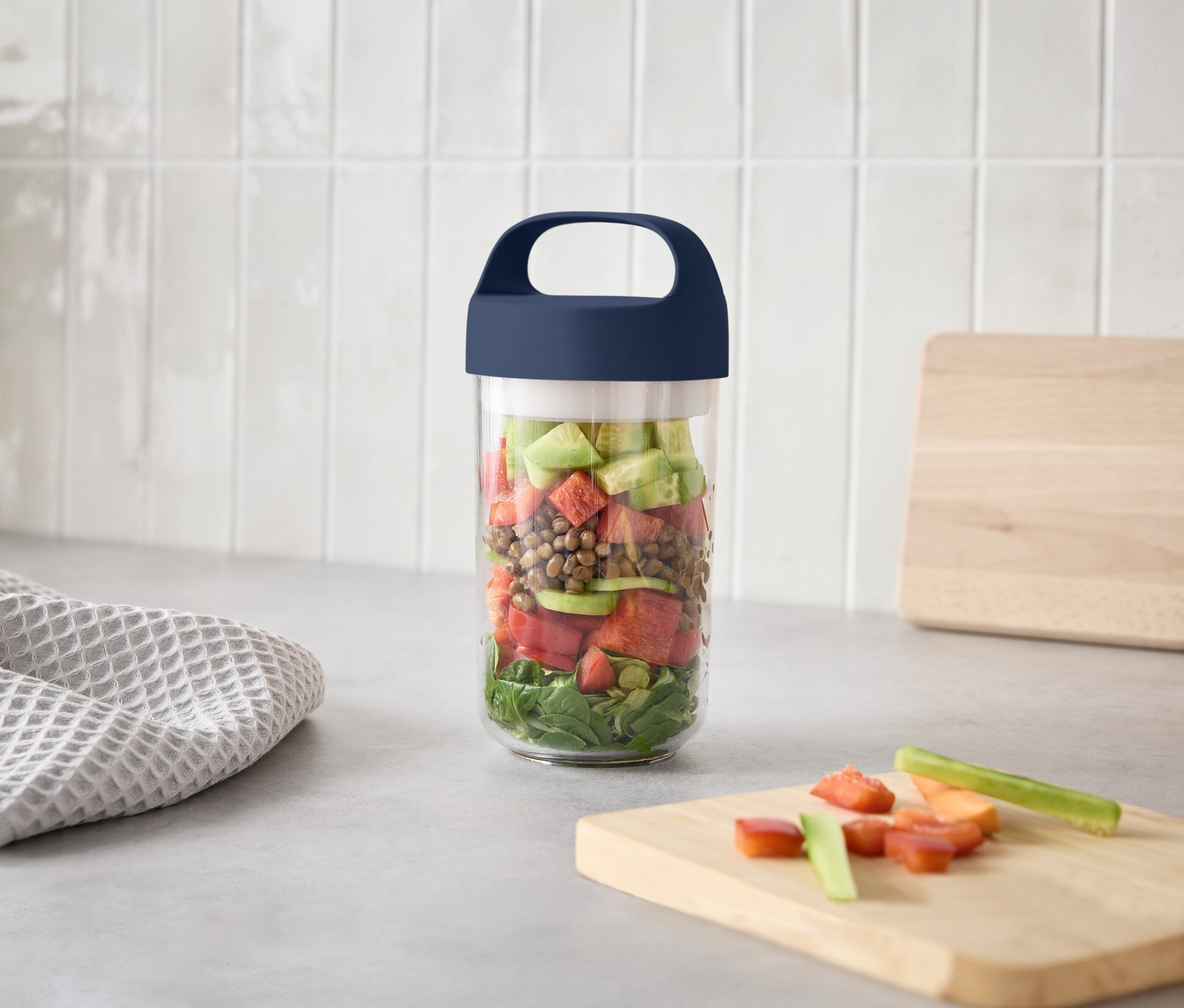 Ein Glas mit Schichtsalat steht auf einer Küchentheke neben einem Schneidebrett mit geschnittenem Gemüse.