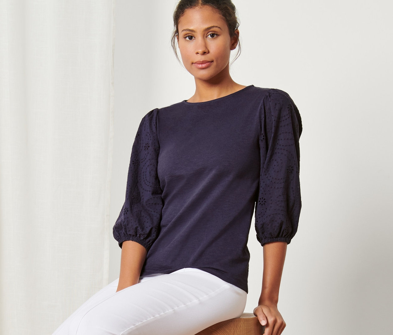 Frau sitzt auf einem Hocker und trägt ein Shirt mit Lochstickerei, weiße Hose und Turnschuhe.