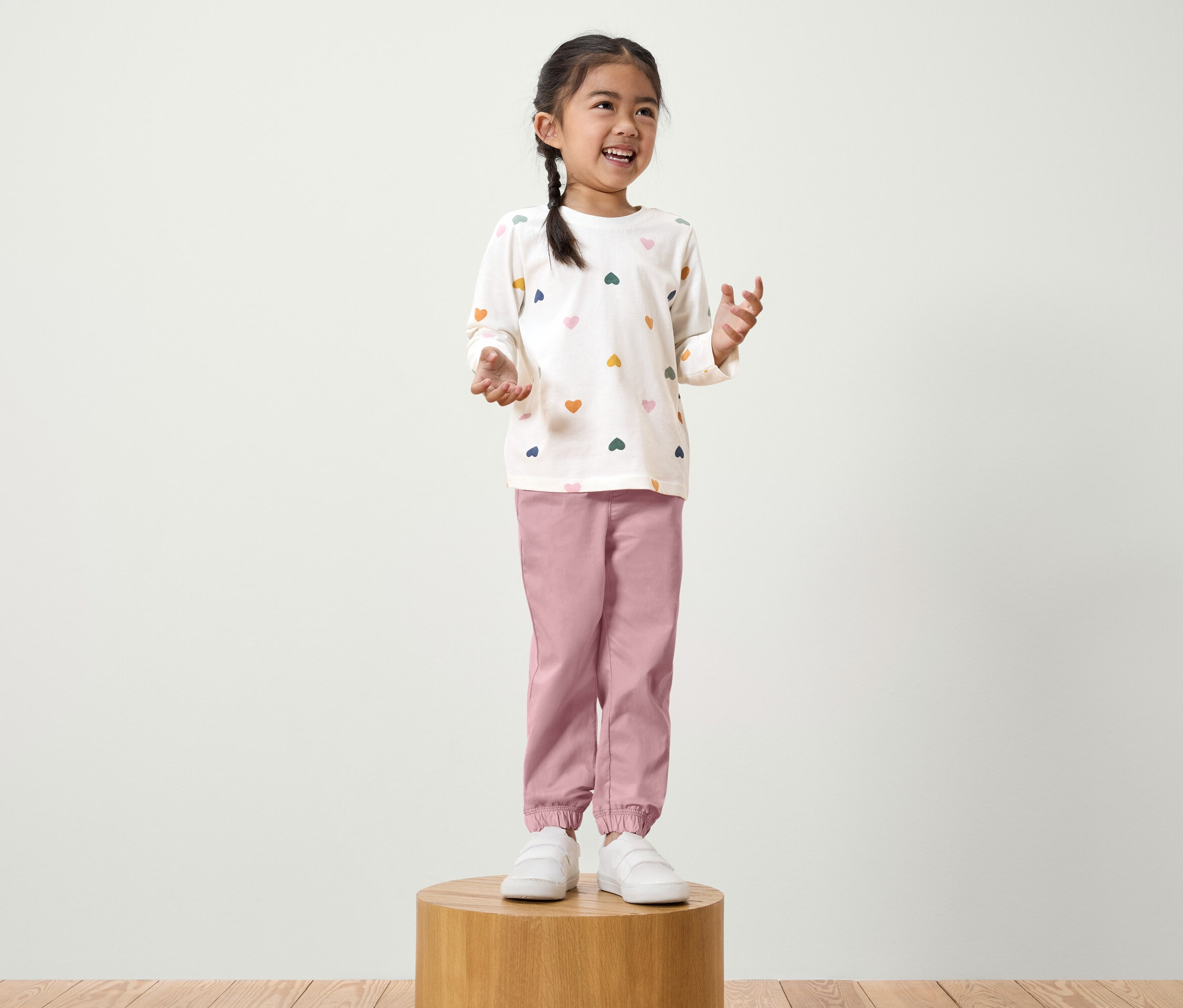 Ein Mädchen mit Zopf steht auf einem Holzsockel. Sie trägt ein weißes Langarmshirt mit bunten Herzen und eine rosafarbene Hose.