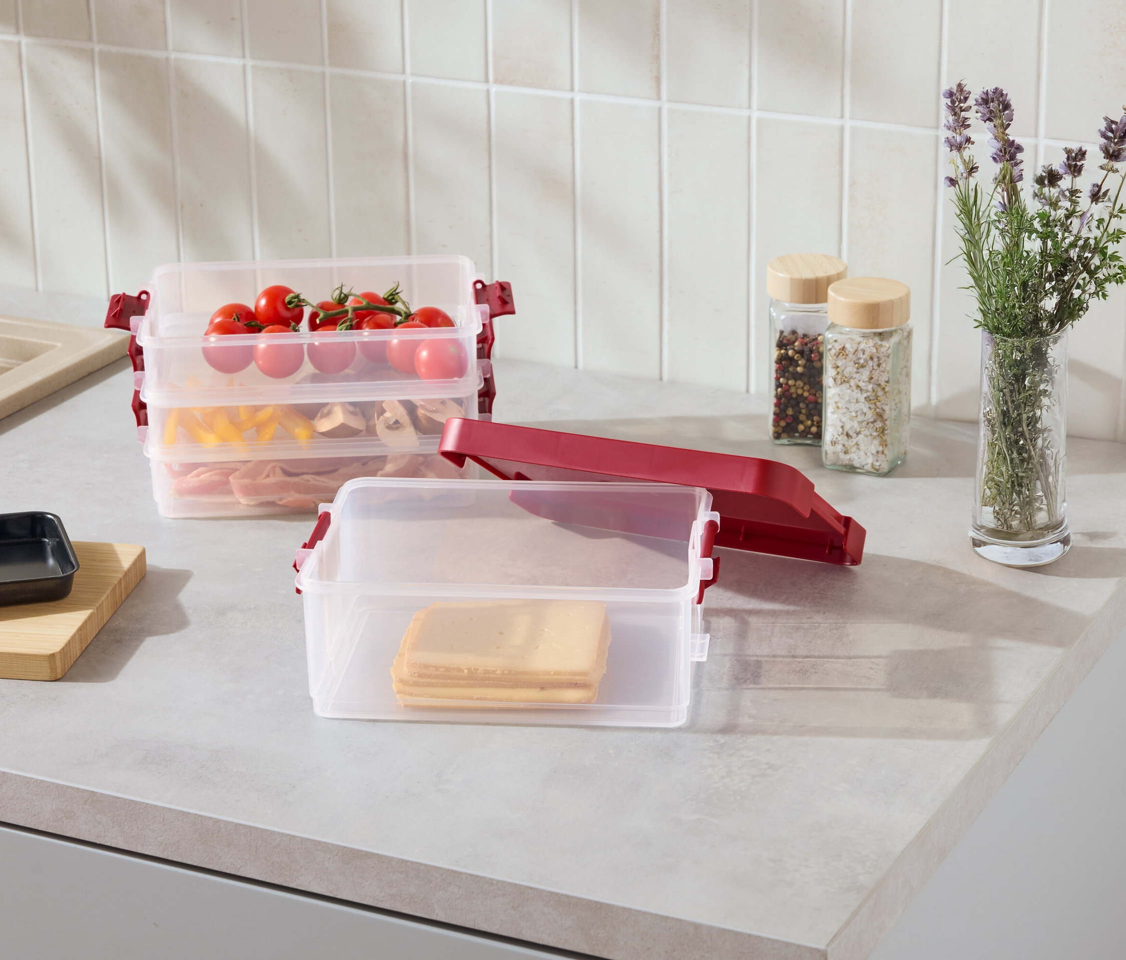 Auf einer Küchentheke stehen durchsichtige, stapelbare Behälter mit roten Clips, gefüllt mit Käsescheiben, Nudeln, Fleisch und Tomaten.
