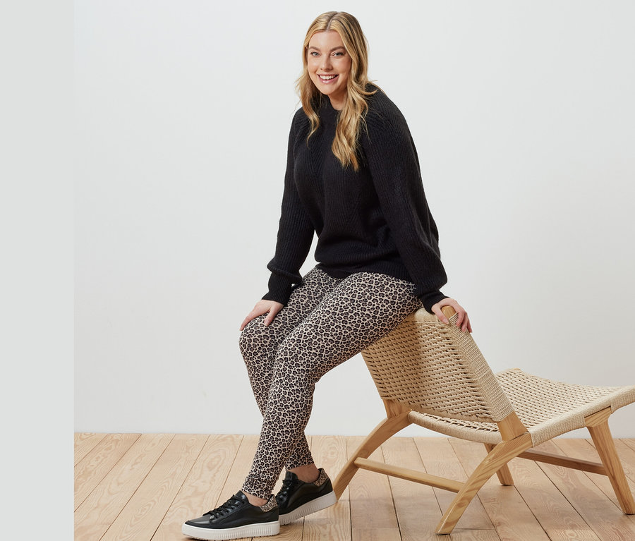 Eine Frau sitzt auf einem Korbsessel und trägt einen schwarzen Strickpullover und bedruckte Leggings.