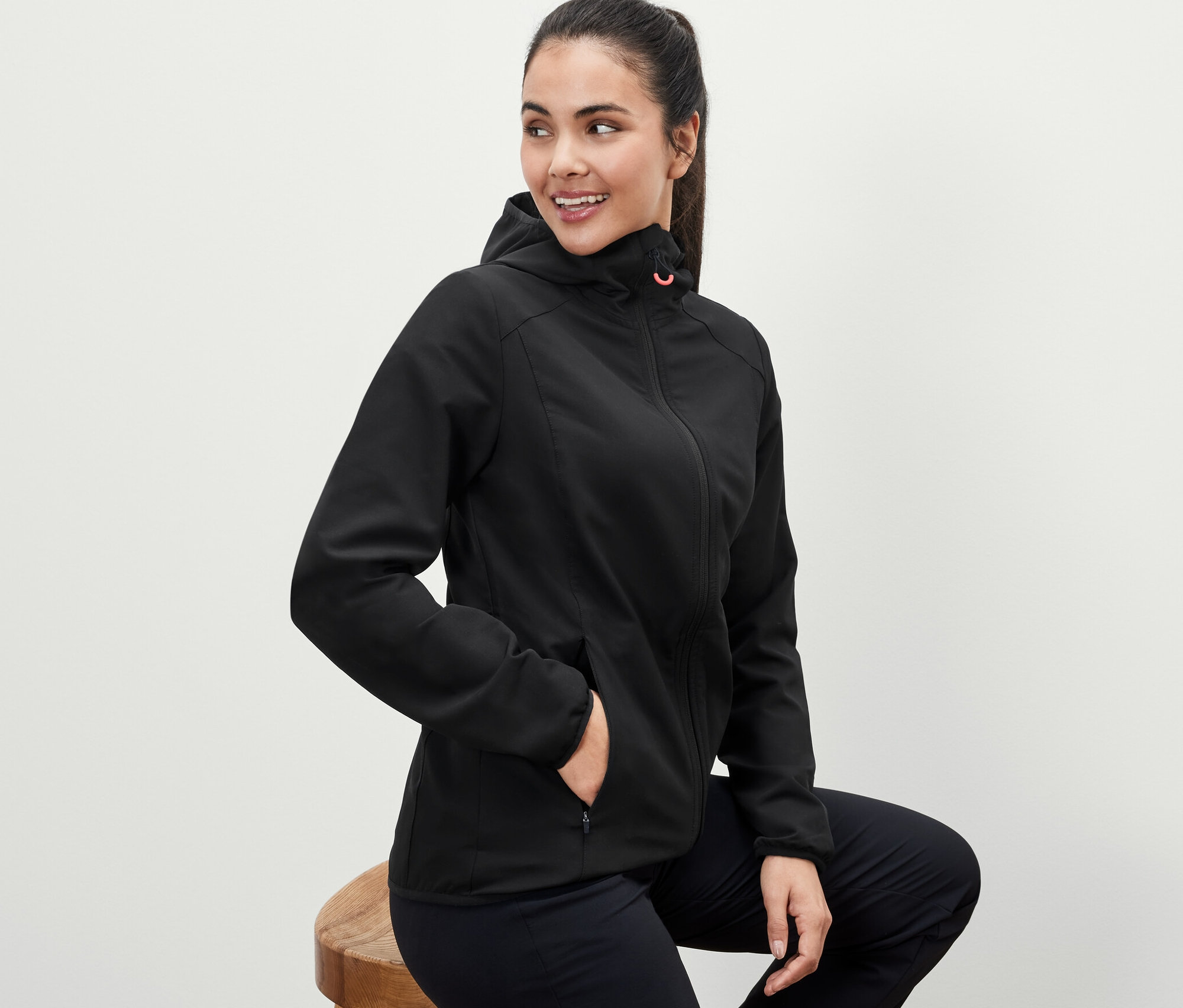 Frau sitzt auf einem Hocker und trägt eine schwarze Softshelljacke.