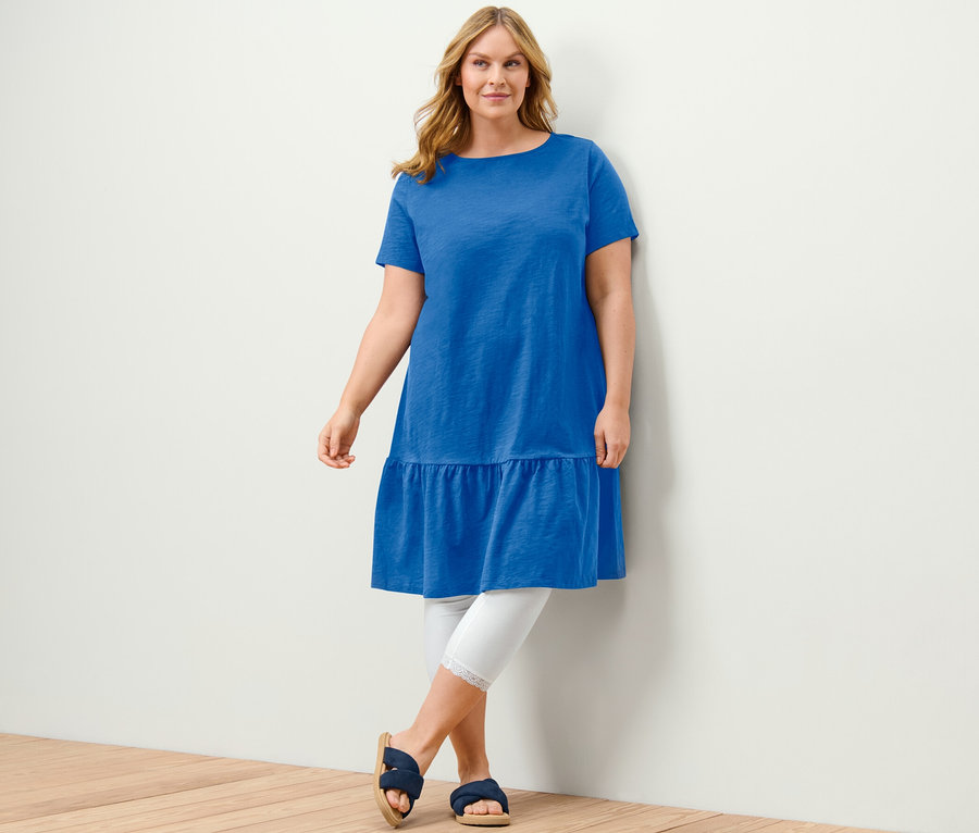 Frau posiert in kobaltblauem Jerseykleid mit Volant, weißen Leggings und blauen Sandalen.