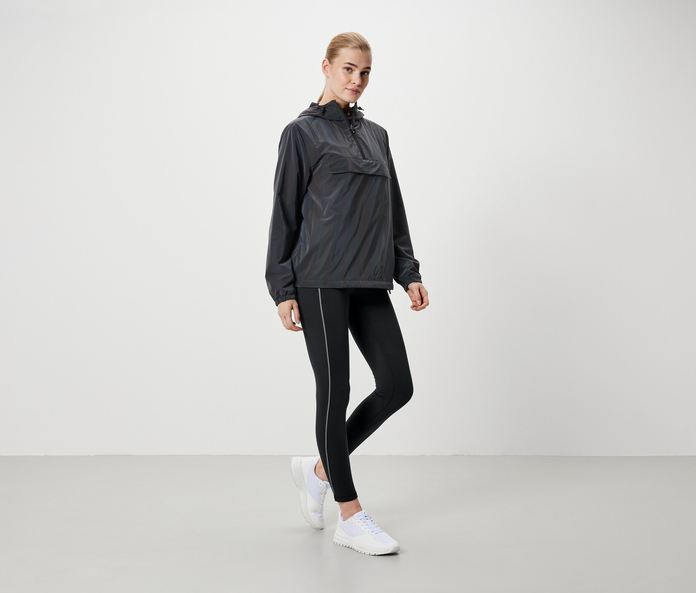 Blonde Frau steht in grauer Kapuzenjacke, schwarzen Leggings und weißen Schuhen.