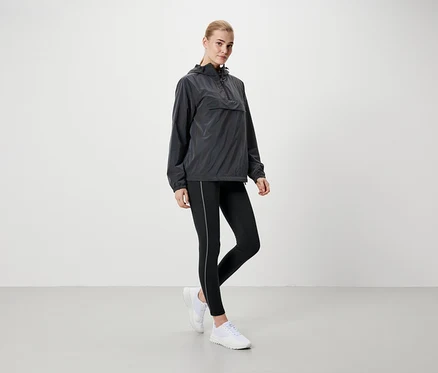 Blonde Frau steht in grauer Kapuzenjacke, schwarzen Leggings und weißen Schuhen.