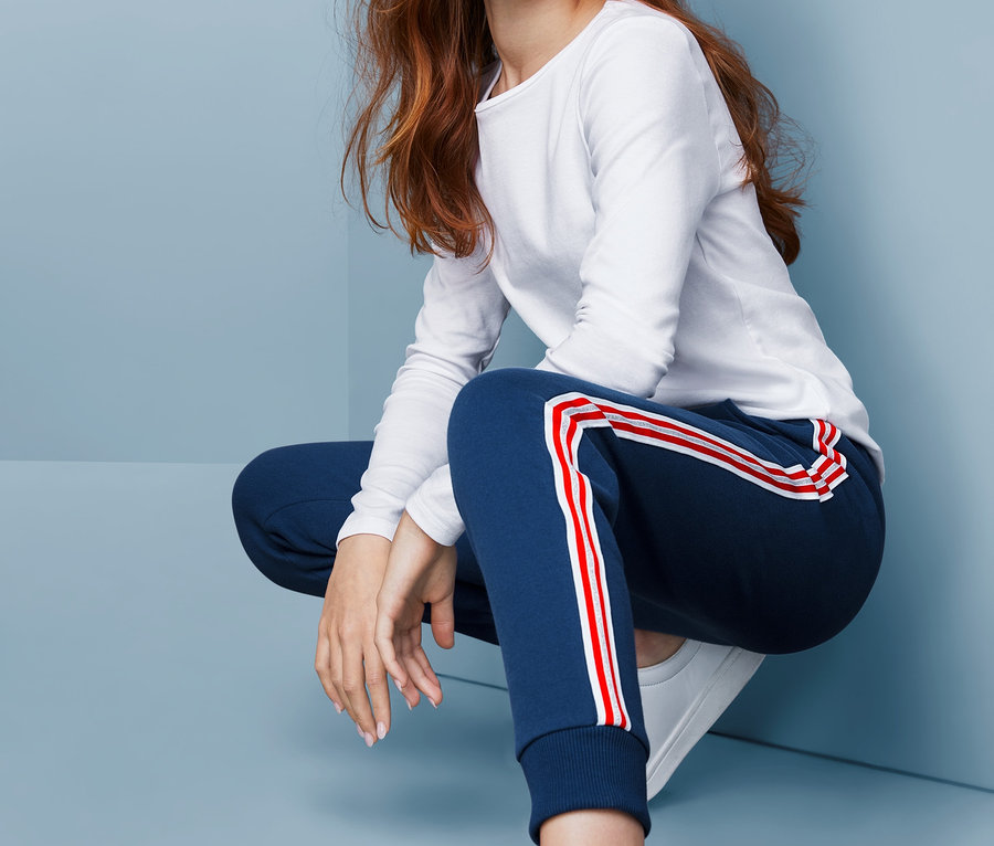 Mädchen in Sweathose mit rot-weißen Streifen, weißem Shirt und weißen Schuhen sitzt vor blauem Hintergrund.