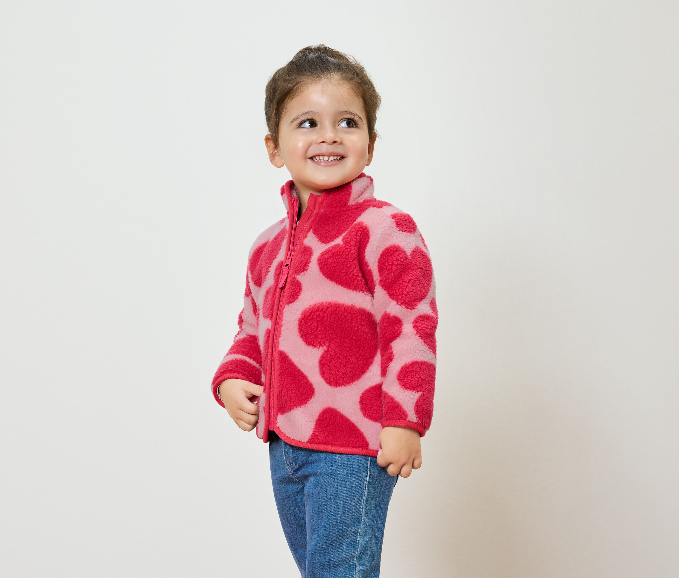Mädchen trägt eine rosa Fleecejacke mit Herzmuster und blaue Jeans, lächelt und schaut zur Seite.