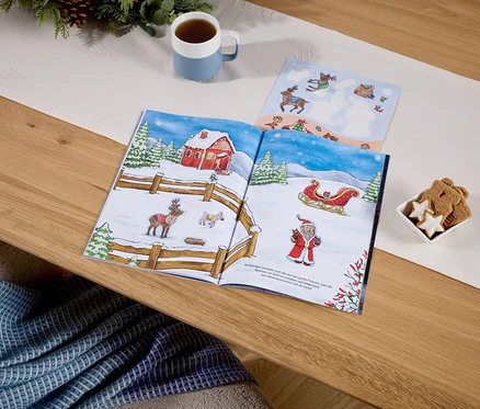 Auf einem Tisch liegt ein aufgeschlagenes Buch mit Weihnachtsbildern, daneben eine Tasse mit Getränk und eine Schale mit Keksen.