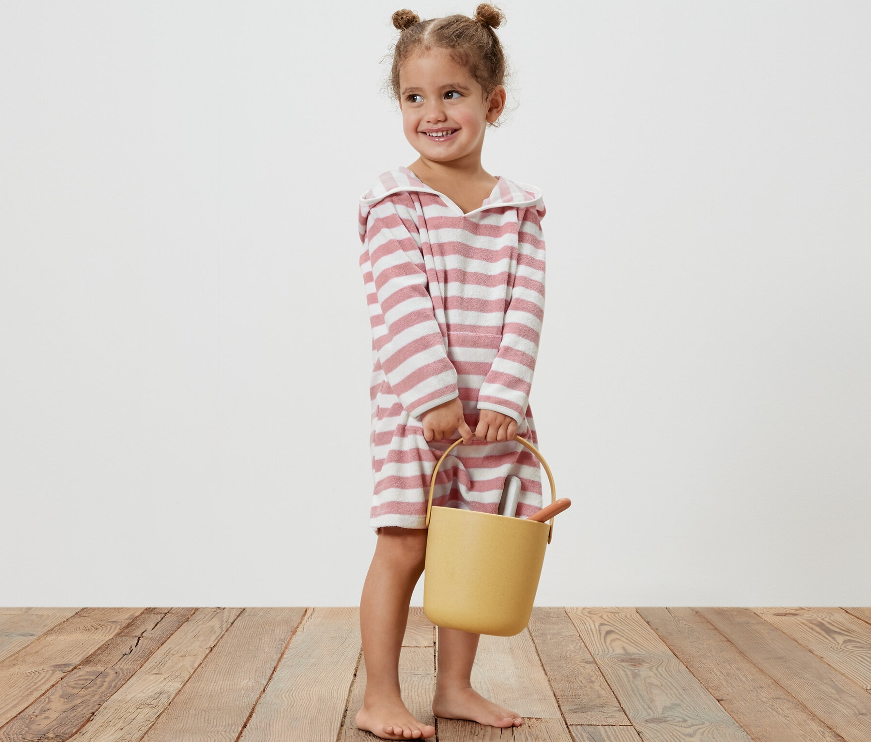 Lächelndes kleines Mädchen hält einen gelben Eimer und trägt einen Kinder-Frottierponcho mit rosa Streifen.