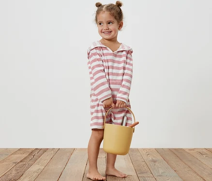 Lächelndes kleines Mädchen hält einen gelben Eimer und trägt einen Kinder-Frottierponcho mit rosa Streifen.