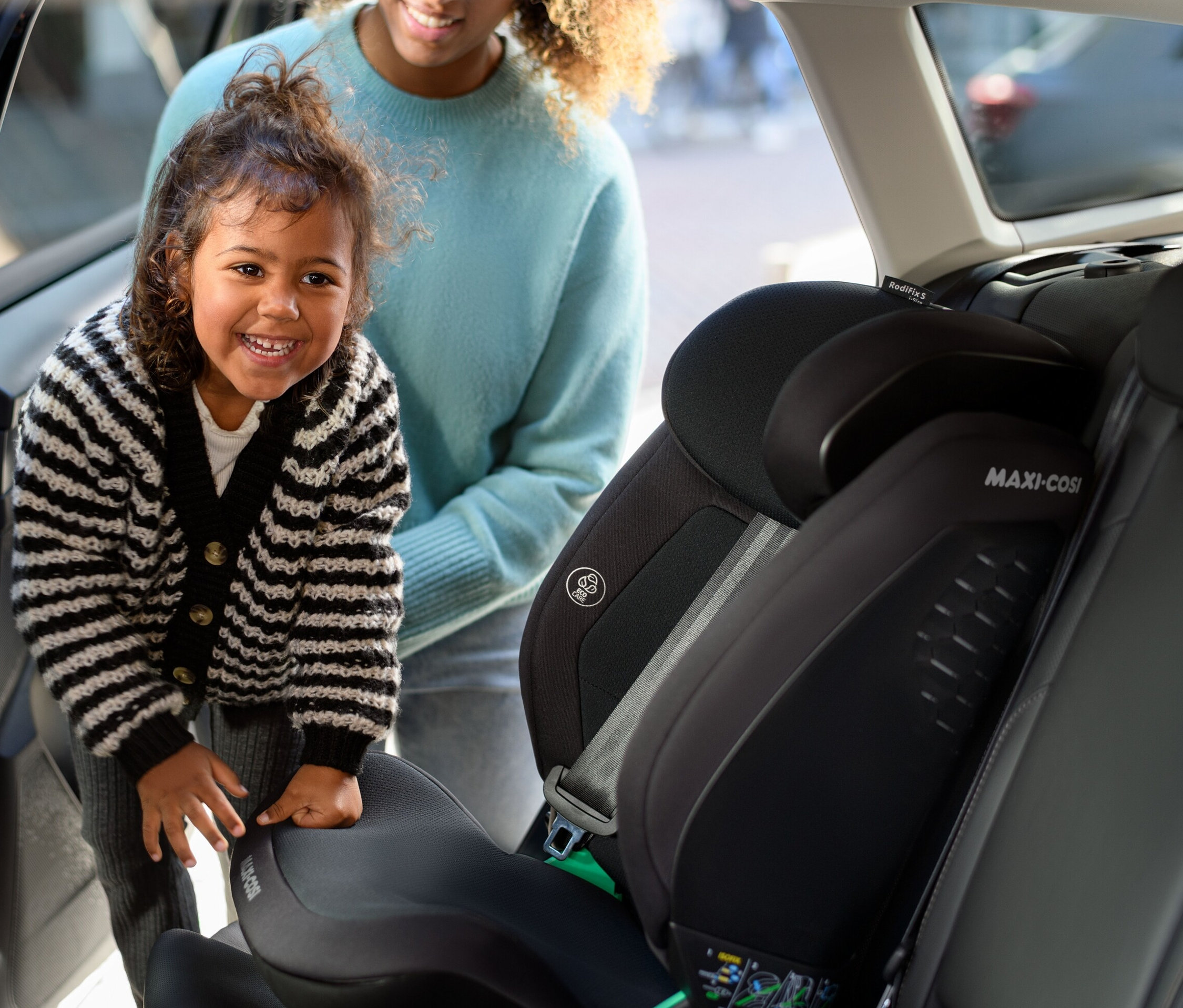 Ein Mädchen lächelt und steht neben einem Maxi-Cosi Kinder-Autositz »Rodifix S«, i-Size im Auto, dahinter steht eine Frau.