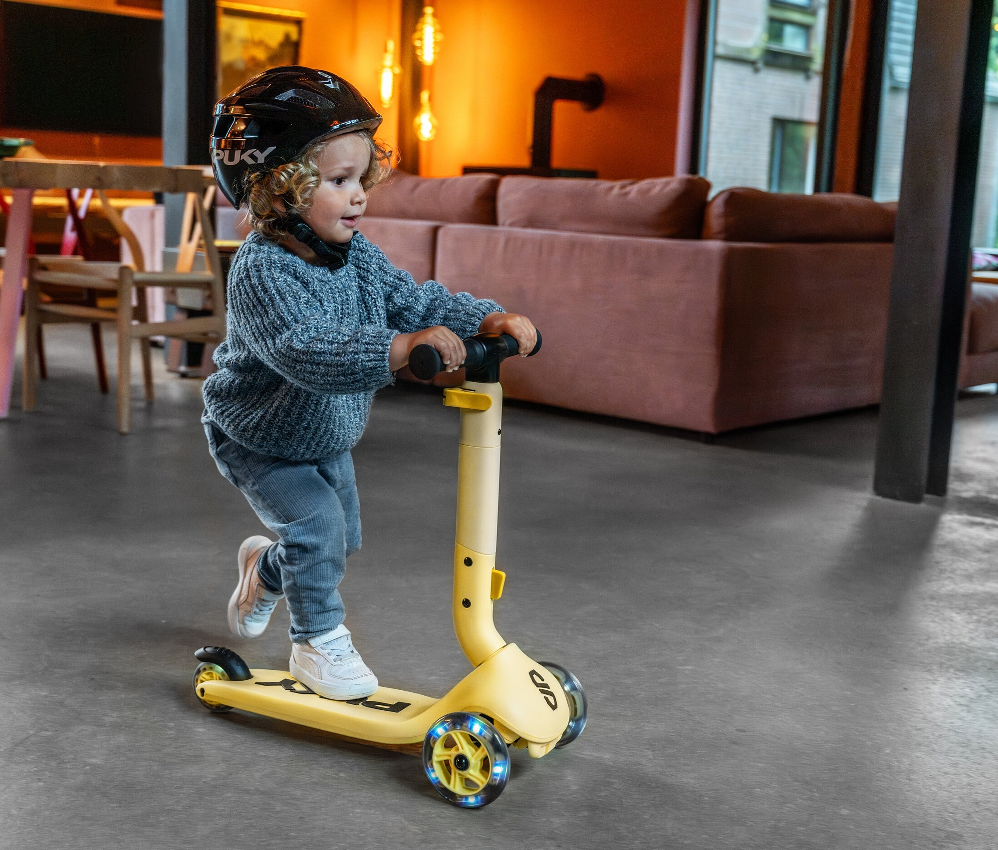 Ein Kind mit Helm fährt mit einem gelben PUKY Roller PUSH im Wohnzimmer.