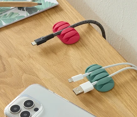 Auf einem Tisch liegen zwei Universal-Kabelmanager, einer in Pink und einer in Grün, zusammen mit einem Telefon und Stiften.