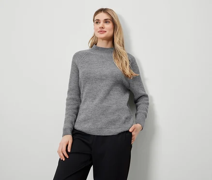 Blonde Frau posiert in grauem Pullover und schwarzer Hose.