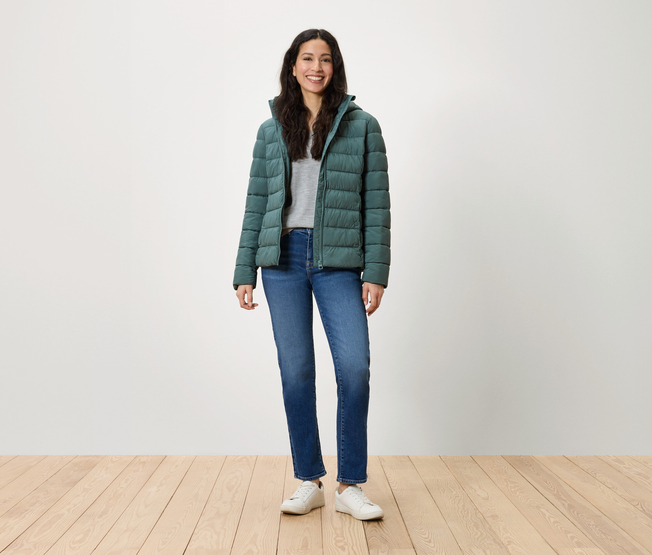 Eine Frau steht und lächelt in einer grünen Daunenjacke, blauen Jeans und weißen Turnschuhen.