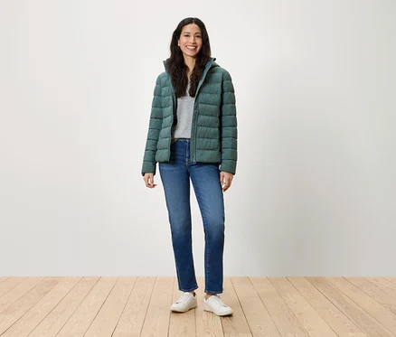 Eine Frau steht und lächelt in einer grünen Daunenjacke, blauen Jeans und weißen Turnschuhen.