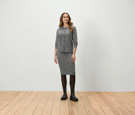Eine Frau steht und trägt eine graue Strickjacke, einen Rock, eine schwarze Strumpfhose und Stiefel.