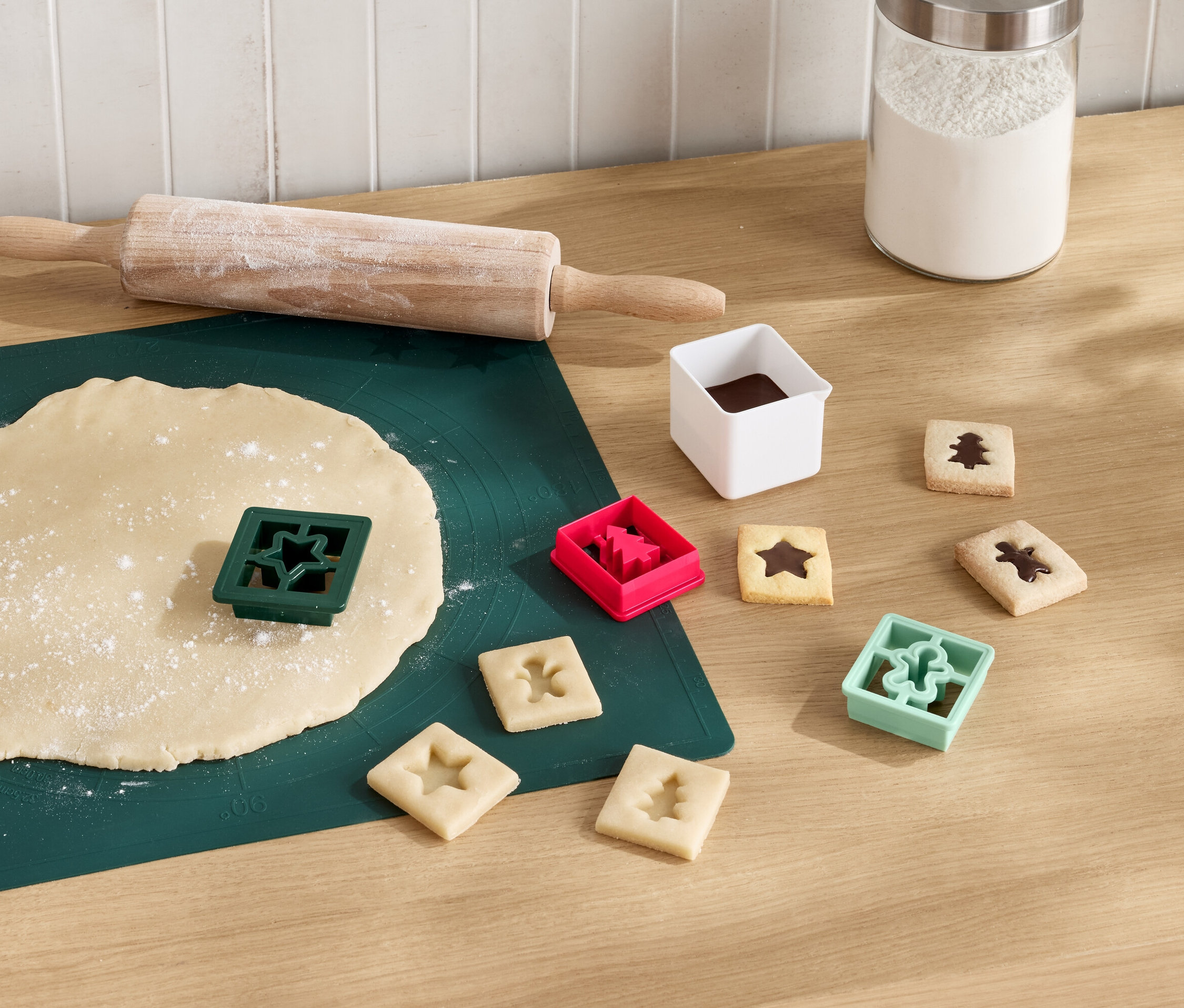 Grüne Unterlage mit ausgerolltem Teig, Nudelholz und sternförmigem Ausstecher. Daneben liegen ausgestochene Kekse mit Schokolade und Ausstecher in Form von Baum, Stern und Männchen.