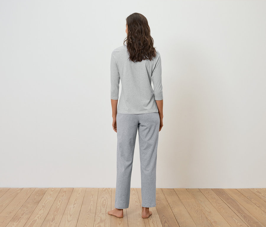 Rückansicht einer Frau mit braunem, gewelltem Haar, die ein graues Dreiviertelarm-Shirt und eine graue Hose trägt.