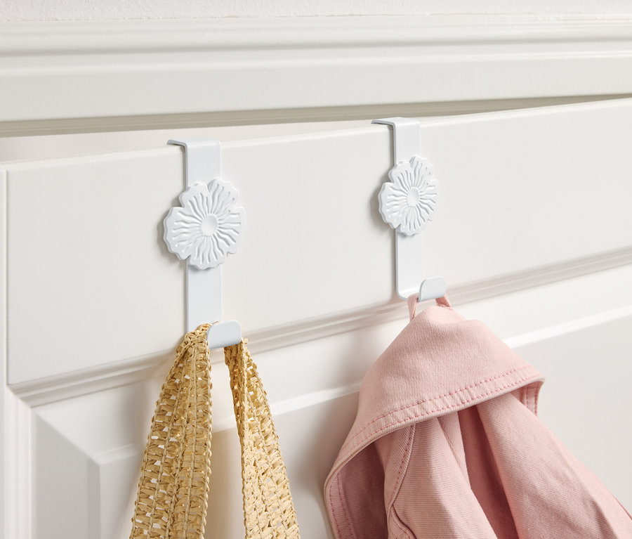Zwei weiße Haken mit Blumendesign hängen an einer weißen Tür, an denen eine gelbe Tasche und eine rosa Jacke hängen.