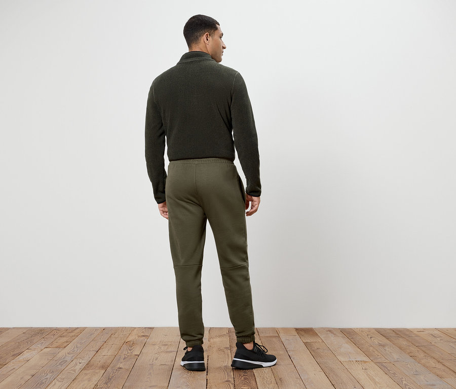 Mann in olivgrünem Fleecepullover und Jogginghose steht mit dem Rücken zur Kamera.