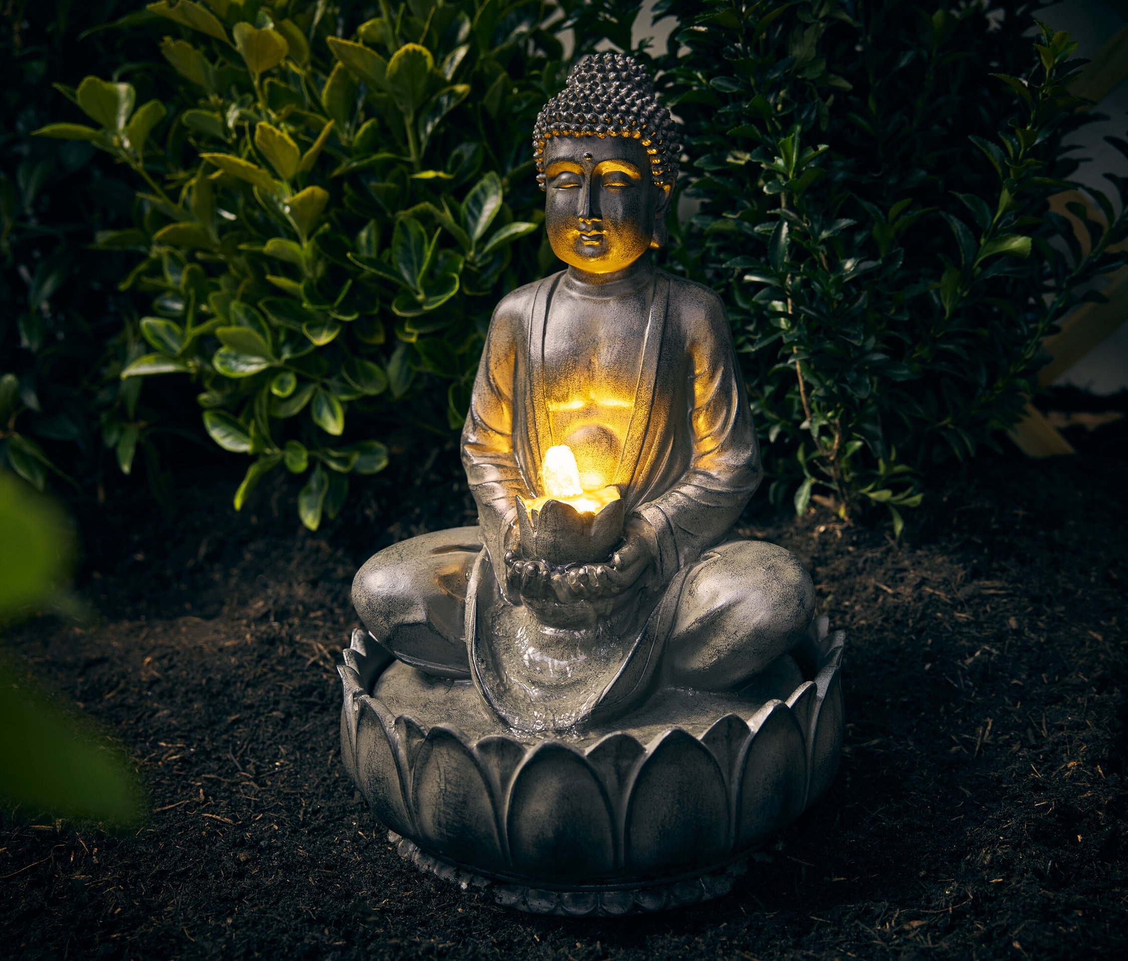 Buddha-Gartenbrunnen sitzt vor einem Busch auf dem Boden.