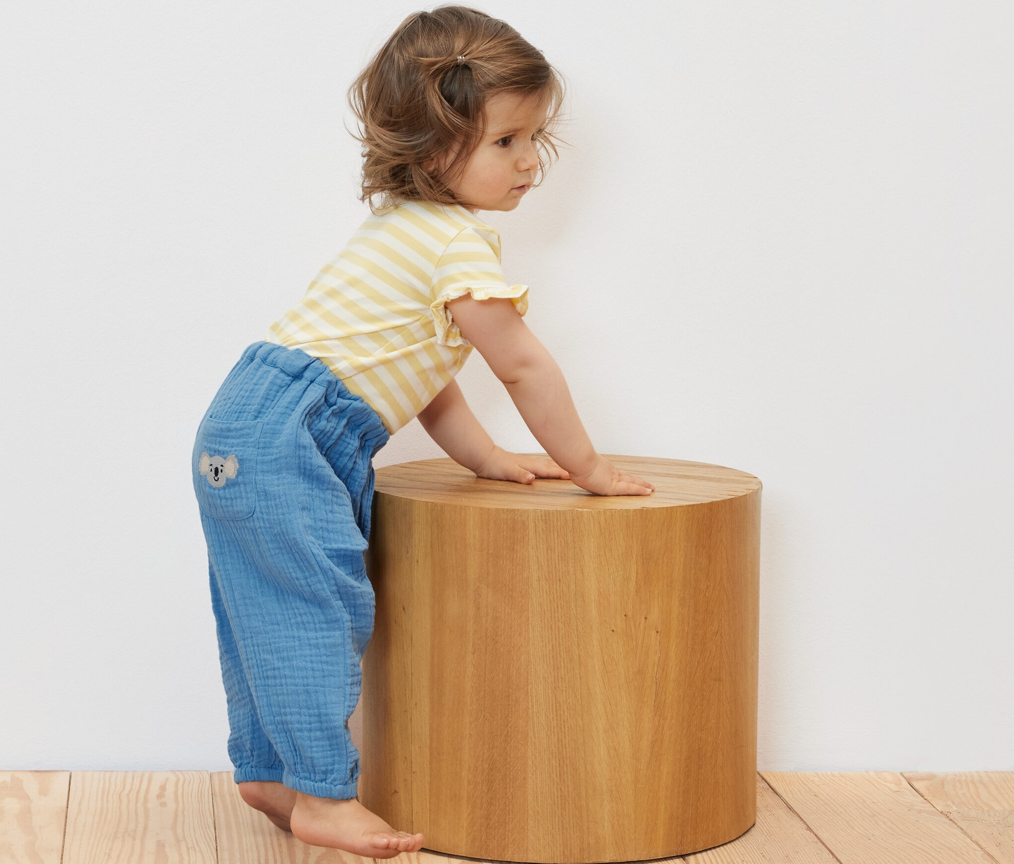 Kleinkind lehnt in Baby-Musselin-Schlupfhose an einem Holzhocker.