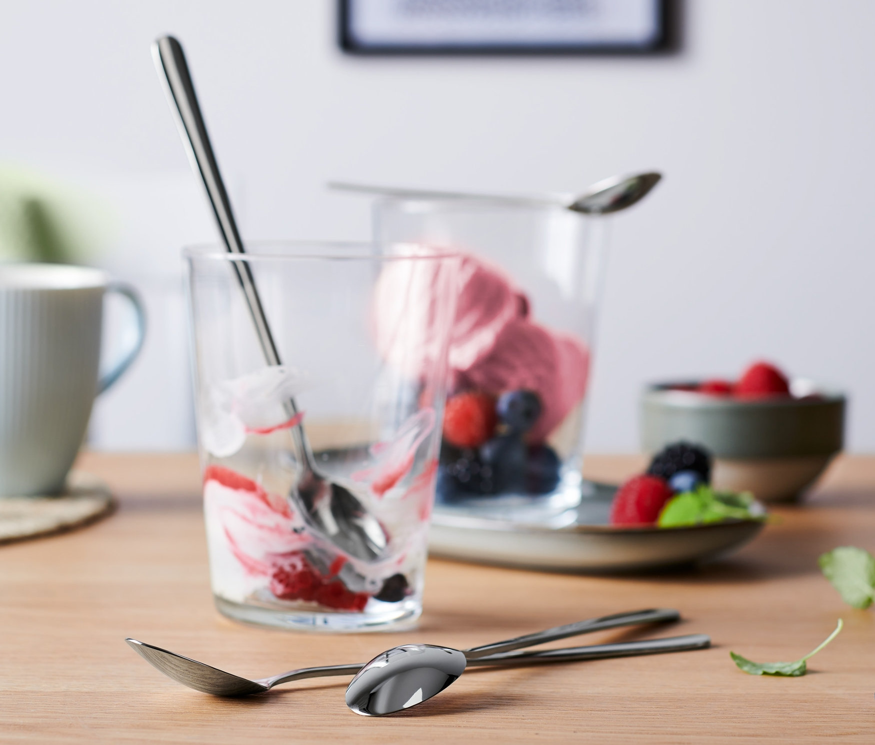 Auf einem Tisch stehen zwei Gläser mit Dessertresten und drei 4 lange Löffel. Im Hintergrund eine Tasse und eine Schale mit Früchten.