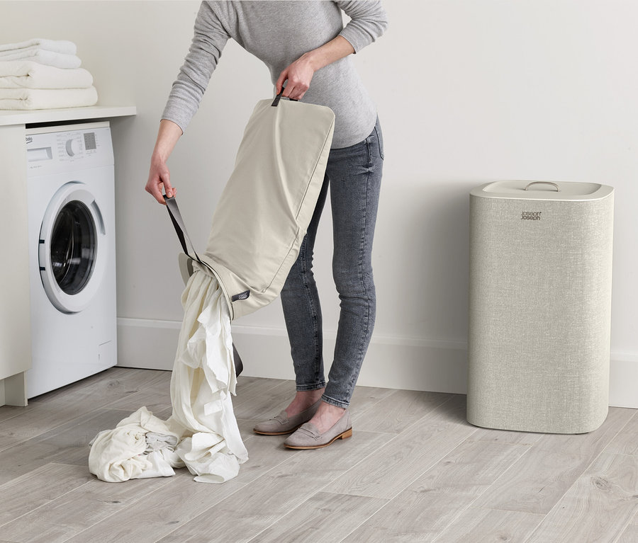 Frau schüttet Wäsche aus einem beigen Wäschesack vor einer weißen Waschmaschine und neben einem beigen Wäschekorb.
