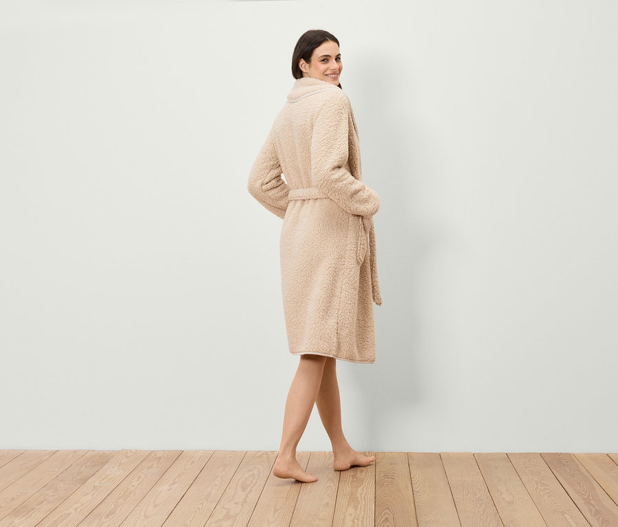 Eine Frau blickt über die Schulter und trägt einen Bademantel aus Kunstfell mit Gürtel, während sie auf einem Holzboden steht.