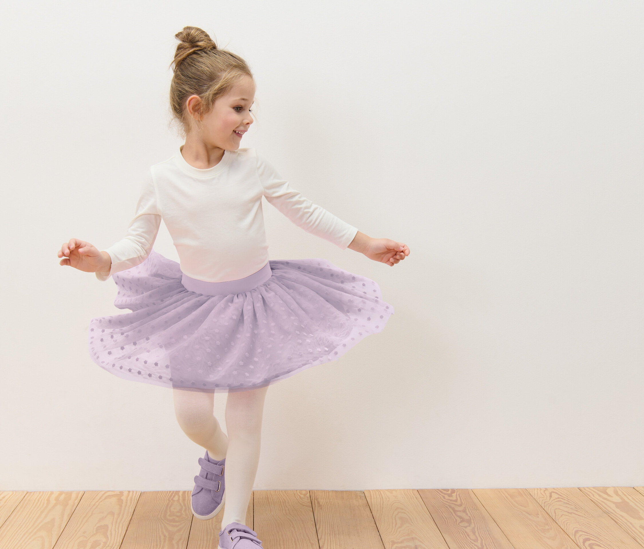 Mädchen mit Dutt tanzt in einem lila Tüllrock mit weißen Punkten, weißem Langarmshirt, Strumpfhose und lila Schuhen vor einer weißen Wand.