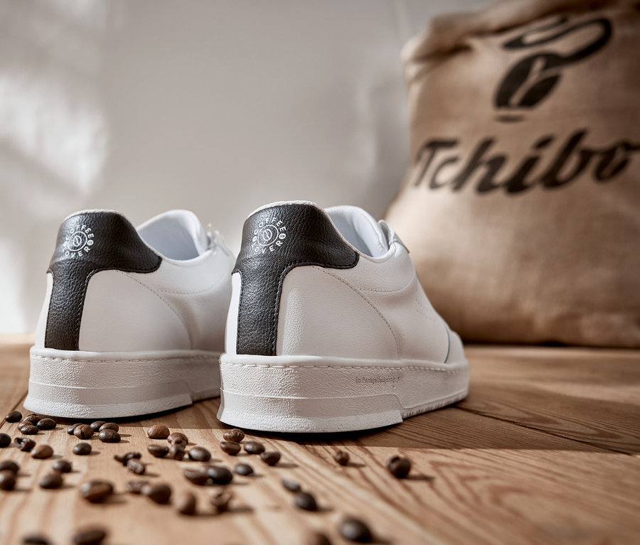 Ein Paar weiße Tchibo x VLACE Coffee Lover Sneaker auf einem Holzboden mit Kaffeebohnen und einem Tchibo Kaffeesack im Hintergrund.