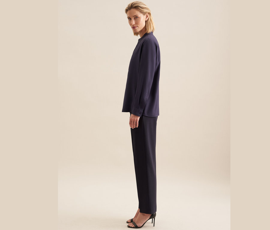 Eine Frau steht seitlich und trägt eine Seidensticker Tunika aus Viskose, eine schwarze Hose und schwarze High-Heel-Schuhe.