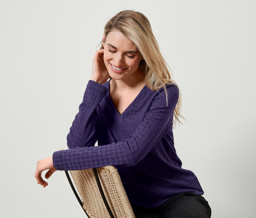 Blonde Frau sitzt auf einem Rattanstuhl, trägt ein violettes Langarmshirt und lächelt.