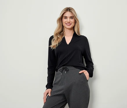 Blonde Frau in schwarzem Langarmshirt und grauer Hahnentrittmusterhose posiert.