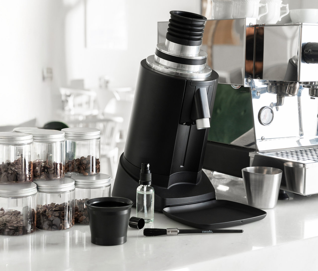 Eine schwarze Kaffeemühle steht auf einer Theke neben Gläsern mit Kaffeebohnen, einem Reinigungsset und einer Espressomaschine.
