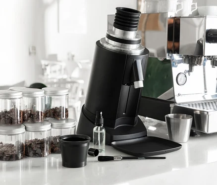 Eine schwarze Kaffeemühle steht auf einer Theke neben Gläsern mit Kaffeebohnen, einem Reinigungsset und einer Espressomaschine.