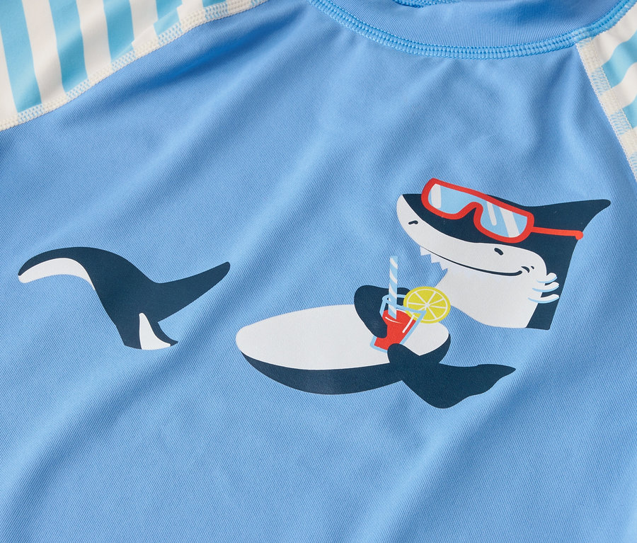 Auf einem hellblauen Stoff ist ein Hai mit Sonnenbrille abgebildet, der an einem Getränk mit Zitrone nippt.