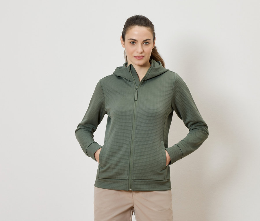 Frau mit olivgrünem Kapuzenpullover mit Reißverschluss und beigen Hosen, die mit beiden Händen in den Taschen steht.