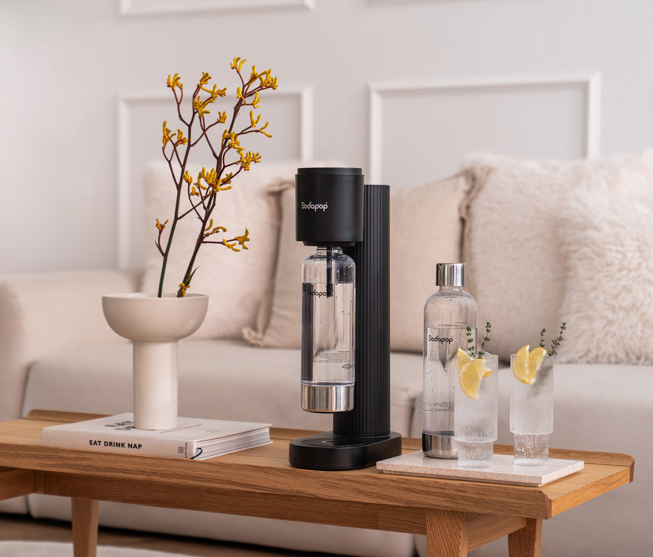 Auf einem Holztisch steht ein Sodapop Wassersprudler Cooper Pro in Schwarz, inklusive einer eingesetzten Flasche. Daneben stehen zwei Gläser mit Zitronenscheiben und eine Sodapop-Flasche.