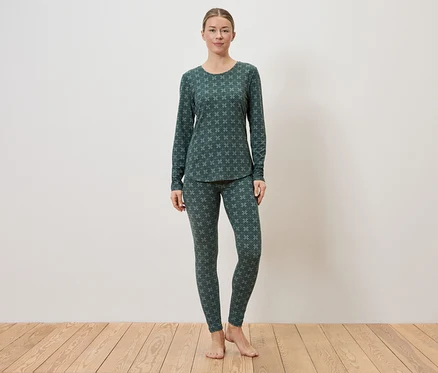 Eine Frau steht barfuß vor einer weißen Wand auf einem Holzboden und trägt einen dunkelgrünen Pyjama mit einem hellgrünen Muster.