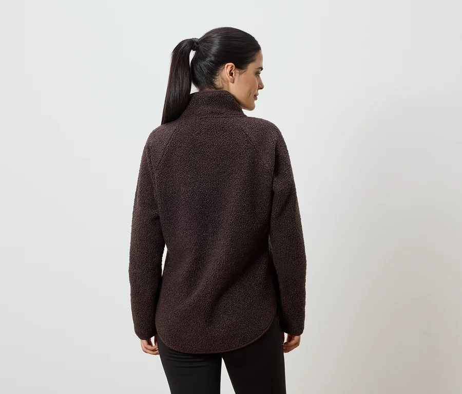 Rückansicht einer Frau mit Pferdeschwanz, die einen braunen Fleecepullover und schwarze Hose vor einem weißen Hintergrund trägt.